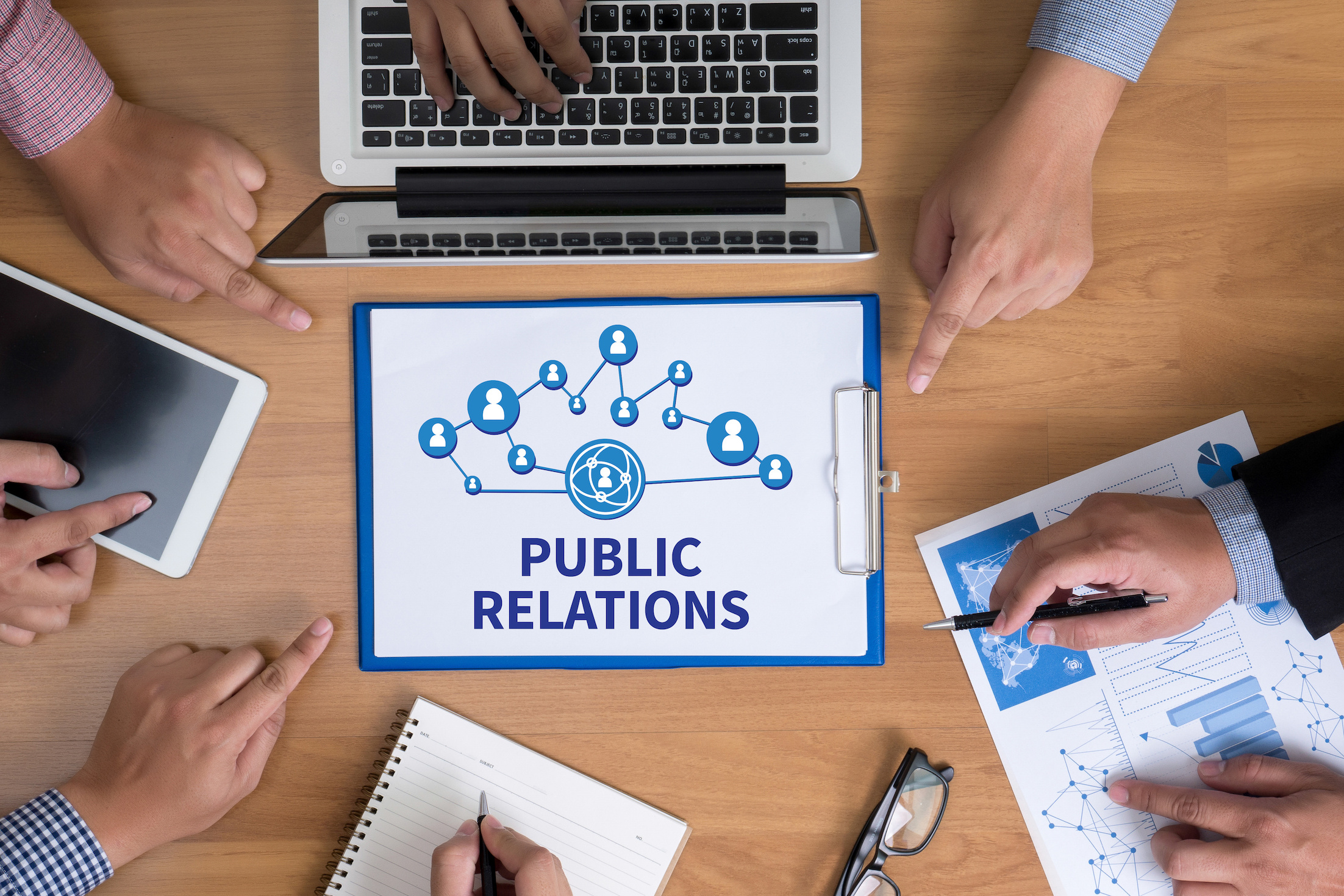 A team works at a table with a laptop. Graphic in the center says public relations.